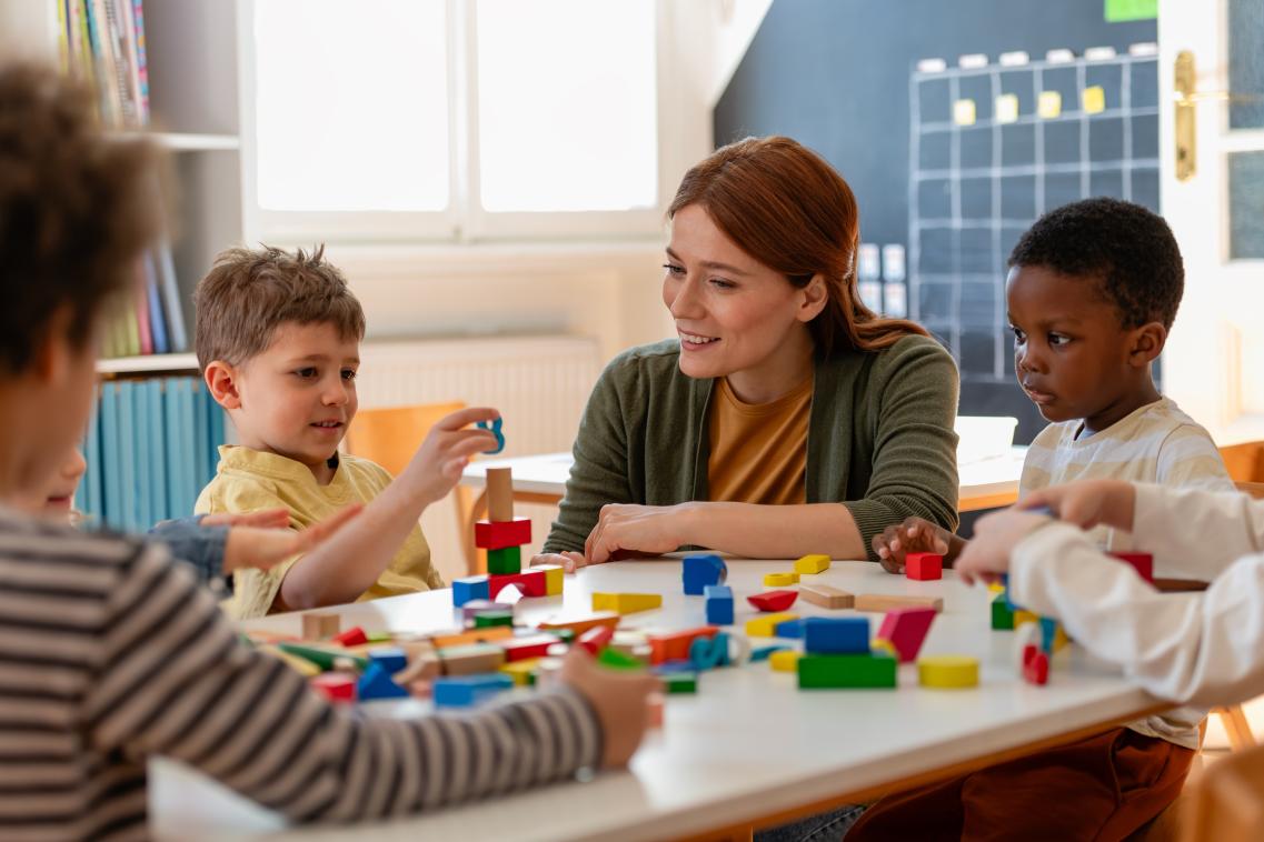 Pre-school children sitting at a table playing with building blocks with a teacher